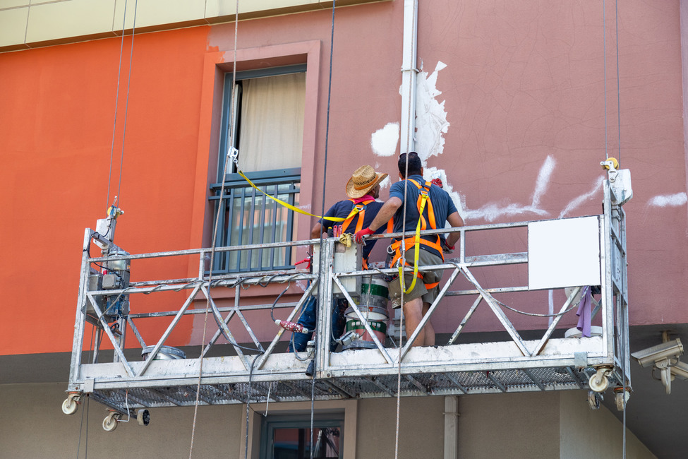 Two plasterers painters performing painting the facade of the house staying in the building cradle. A view of the facade from the back of workers, restoration process at height in lifting bucket